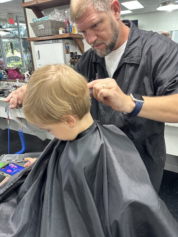 A kid getting a haircut, cape on, scissor work up top. Garner, NC, 2025.